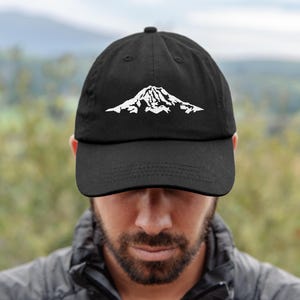May include: Black baseball cap featuring a white mountain range graphic. The cap has a curved brim and an adjustable strap. The mountain design is a simple, stylized outline. This cap is suitable for outdoor activities or casual wear.