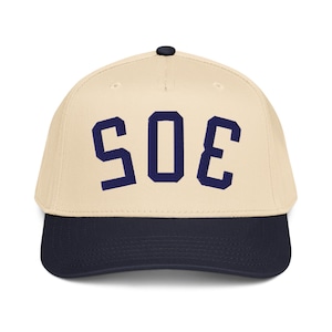 Pode incluir: Boné de beisebol com coroa creme e aba azul marinho. O boné apresenta o número "305" bordado em letras de bloco azul marinho na frente. O boné tem um botão preto na parte superior.