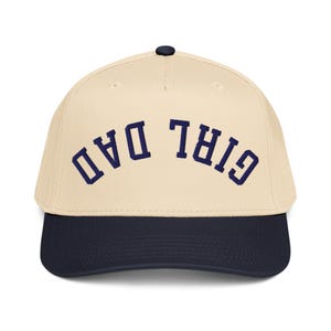 May include: A baseball cap with a cream-colored crown and a navy blue brim. The text "GIRL DAD" is embroidered in navy blue across the front of the cap. The cap has a black button on top.