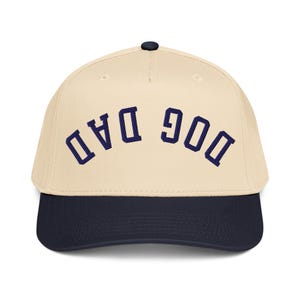 May include: A baseball cap in cream and navy blue. The cream-colored crown features the navy blue embroidered text "DOG DAD" in an arch. The cap has a navy blue brim and a black button.
