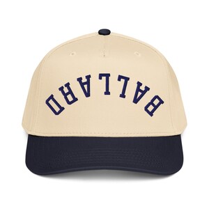May include: A cream-colored baseball cap with a navy blue brim and a navy blue button on top. The word "BALLARD" is embroidered in navy blue across the front of the cap.