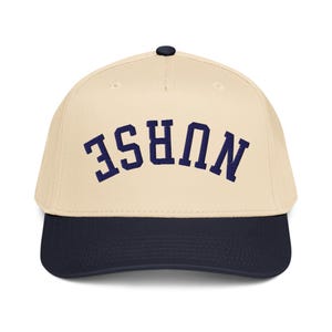 May include: A baseball cap with a cream-colored crown and a navy blue brim. The word "NURSE" is embroidered in navy blue across the front. The cap has a curved bill and a black button on top.