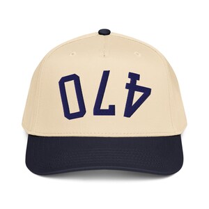 Pode incluir: Boné de beisebol com coroa creme e aba azul marinho. O boné apresenta o número "470" em letras de bloco azul marinho na frente. O boné tem um botão preto na parte superior.