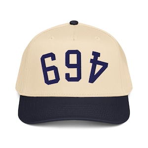 Pode incluir: Boné de beisebol com coroa creme e aba azul marinho. O boné apresenta o número "694" em azul marinho na frente. Possui um botão preto na parte superior.