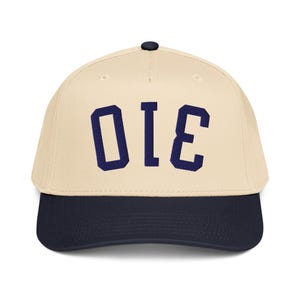 Pode incluir: Boné de beisebol com coroa creme e aba azul marinho. O boné apresenta as letras azul marinho "OIE" na frente. O boné tem um botão preto na parte superior.