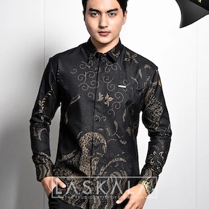 May include: Black button-down shirt with a gold floral pattern. The shirt has long sleeves and a pocket on the chest. The word "LASKAL" is printed on the bottom of the shirt.