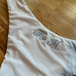 Könnte beinhalten: Ein weißes, asymmetrisches Stoffteil mit dem gestickten Text "Mrs M" in hellblauer Schrift. Der Stoff hat eine glatte Textur. Das Objekt liegt auf einer Holzoberfläche.