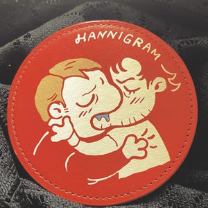 Może przedstawiać: Okrągły, czerwony podkładka pod szklankę z napisem "HANNIGRAM" w stylizowanej czcionce. Podkładka przedstawia kreskówkową ilustrację dwóch postaci w bliskim uścisku, ze złotymi i niebieskimi akcentami. Podkładka jest prawdopodobnie wykonana ze skóry lub podobnego materiału.