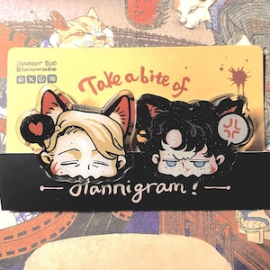Pode incluir: Dois pinos de acrílico transparente com personagens em estilo anime com orelhas de gato. Um tem cabelo loiro e um coração, o outro tem cabelo preto e um rosto zangado. Os pinos estão em um cartão amarelo com o texto "Take a bite of" e "Glannigram!"