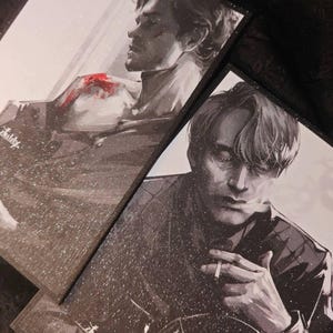Puede incluir: Dos impresiones artísticas enmarcadas con ilustraciones en blanco y negro de hombres. Una muestra a un hombre con una mancha roja, la otra a un hombre fumando. Ambas impresiones tienen un efecto brillante y un estilo dramático.