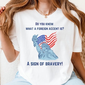 May include: White t-shirt with a blue and red Statue of Liberty graphic and the text "Do you know what a foreign accent is? A sign of bravery!"