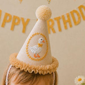 Peut inclure: Un chapeau de fête crème avec un pompon jaune et un bord à volants. Le chapeau présente un motif de canard brodé avec un nœud jaune et des pieds orange. Une bannière avec l'inscription "HAPPY BIRTHDAY" est en arrière-plan.