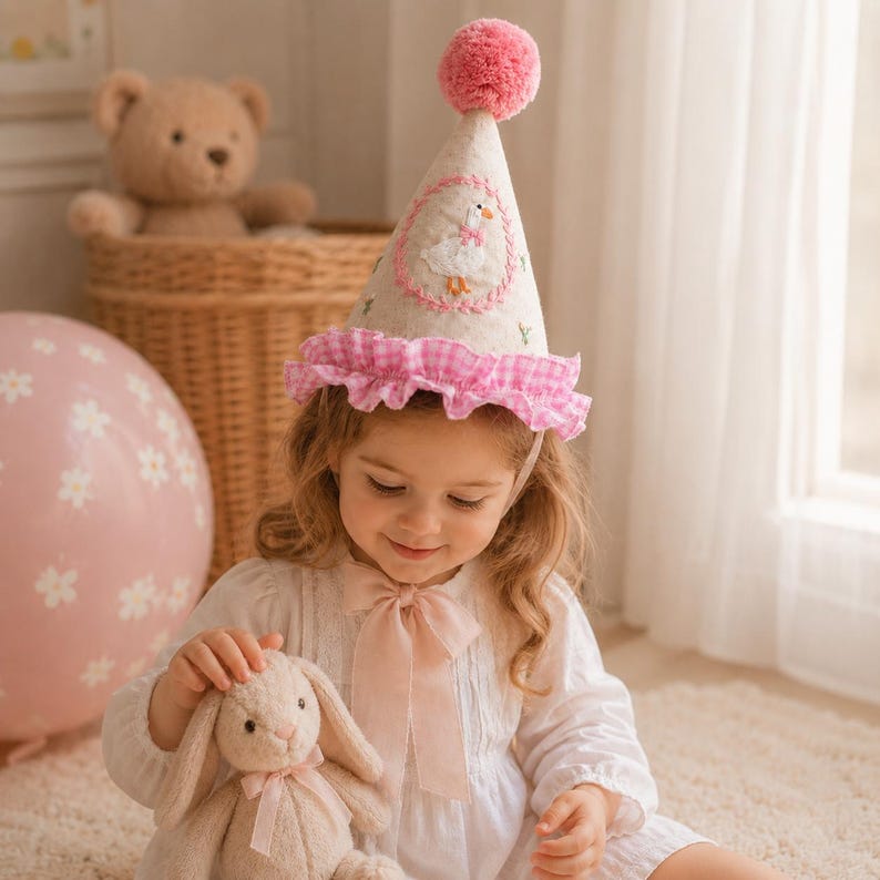 May include: A conical party hat made of beige fabric featuring a pink pom-pom on top, a pink gingham ruffled trim, and an embroidered circular patch depicting a white goose with an orange beak. The hat is secured with a light pink ribbon tie.