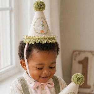 K&ouml;nnte beinhalten: Eine cremefarbene Partyhut mit einem gr&uuml;nen Bommel und einer R&uuml;schenborte. Der Hut zeigt ein gesticktes Enten-Design in einem gr&uuml;nen Kranz. Ein rosa Band sichert den Hut. Der Hut ist f&uuml;r eine Kindergeburtstagsfeier konzipiert.