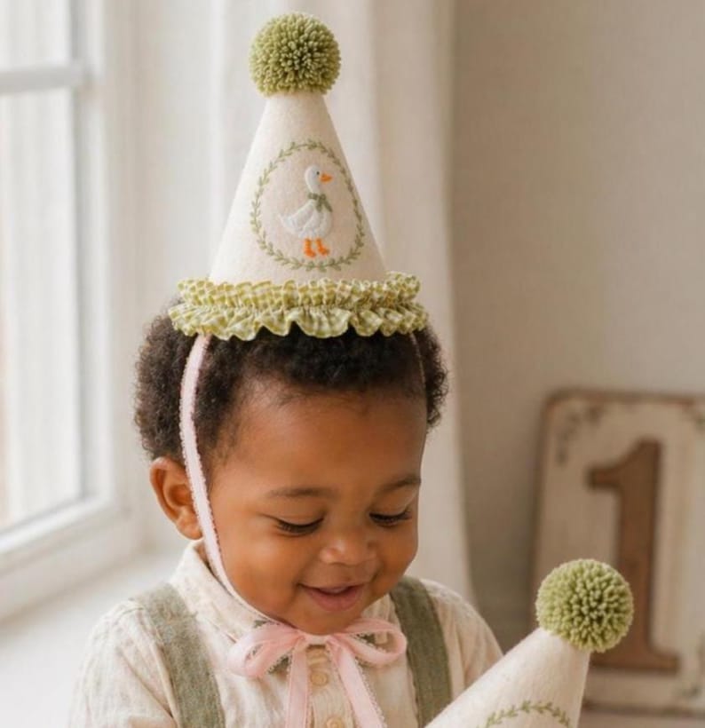 K&ouml;nnte beinhalten: Eine cremefarbene Partyhut mit einem gr&uuml;nen Bommel und einer R&uuml;schenborte. Der Hut zeigt ein gesticktes Enten-Design in einem gr&uuml;nen Kranz. Ein rosa Band sichert den Hut. Der Hut ist f&uuml;r eine Kindergeburtstagsfeier konzipiert.