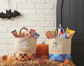 Puede incluir: Dos bolsas de golosinas de Halloween llenas de caramelos, con diseños de fantasmas bordados y nombres personalizados. Una bolsa tiene una base a cuadros naranja y negro, la otra una base a cuadros azul y morado. Una calabaza y una decoración de murciélago completan la escena festiva.