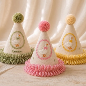 May include: Three cone-shaped party hats in cream felt, each topped with a pom-pom and featuring a ruffled gingham base. The hats display embroidered goose motifs within floral wreaths in green, pink, and yellow, with the name Kenny on one.