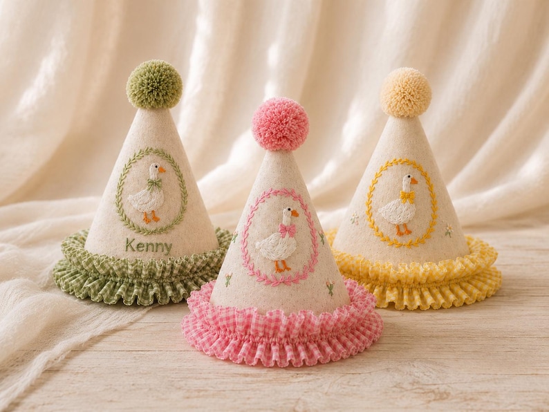May include: Three cone-shaped party hats in cream felt, each topped with a pom-pom and featuring a ruffled gingham base. The hats display embroidered goose motifs within floral wreaths in green, pink, and yellow, with the name Kenny on one.