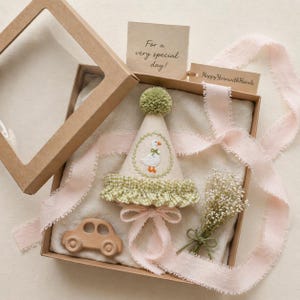 K&ouml;nnte beinhalten: Eine Geschenkbox mit einer Partyhut, einem Holzauto und Trockenblumen. Der Partyhut ist cremefarben mit einem gr&uuml;nen Bommel und einer Entenstickerei. Die Box enth&auml;lt ein Etikett mit "For a very special day!" und ein Band.