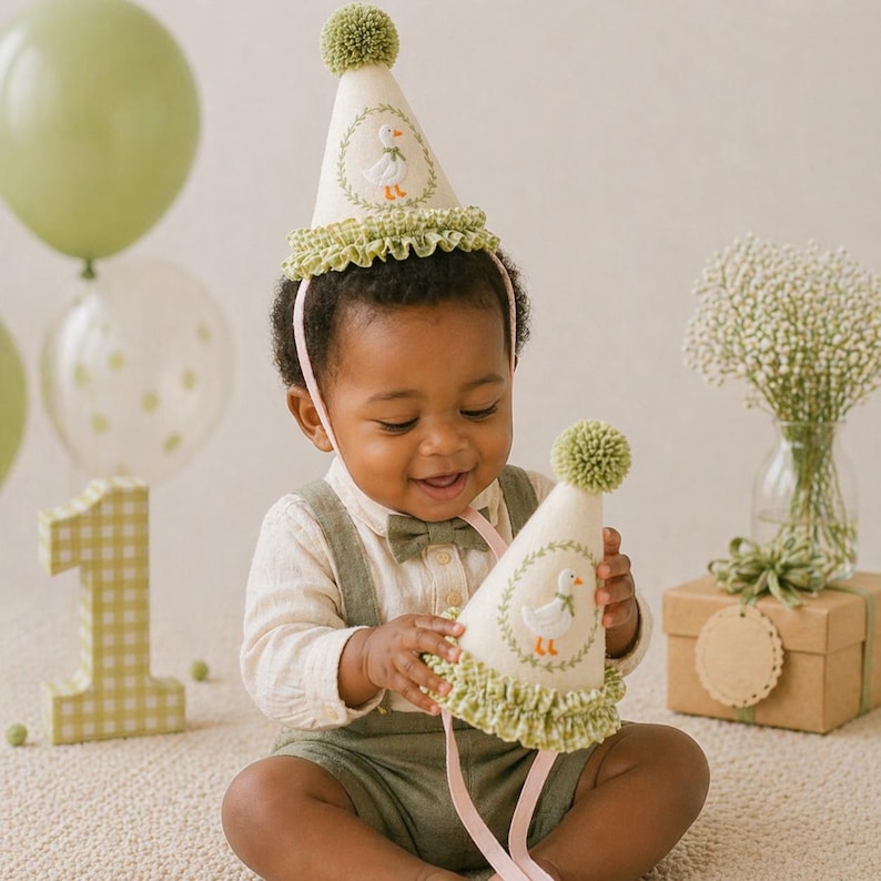 K&ouml;nnte beinhalten: Ein Baby tr&auml;gt einen wei&szlig;en Partyhut mit einem gr&uuml;nen Bommel und einem G&auml;nsemotiv. Der Hut hat eine gr&uuml;ne R&uuml;schenborte und ein rosa Stirnband. Das Baby tr&auml;gt ein helles Hemd, gr&uuml;ne Shorts und eine Fliege. Gr&uuml;ne Luftballons und die Zahl 1 sind im Hintergrund.