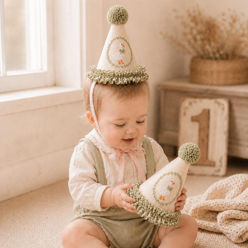 May include: Two cone-shaped party hats made of off-white fabric, each topped with a sage green pom-pom. The hats feature a circular embroidered wreath design with a white goose in the center and a sage green and white gingham ruffled trim.