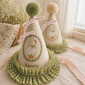 K&ouml;nnte beinhalten: Zwei kegelf&ouml;rmige Partyh&uuml;te in Beige mit einer G&auml;nse-Stickerei. Die H&uuml;te haben gr&uuml;ne Akzente, darunter eine R&uuml;schenborte und ein Pompon. Auf einem Hut ist der Name "Kenny" gestickt. Die H&uuml;te sind mit einer Gans mit Schleife verziert.