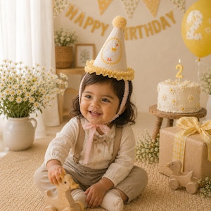 May include: A cream-colored cone-shaped party hat featuring a yellow pom-pom on top, a ruffled yellow trim at the base, and an embroidered white duck with an orange beak and feet. The hat includes a delicate pink lace chin strap for securing.