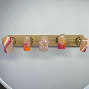 Può includere: Un set di cinque unghie finte con vari design. Le unghie presentano una base nude con accenti arancioni, rosa e bianchi. Un'unghia ha un fiore rosa 3D. Le unghie sono esposte su un supporto di legno.