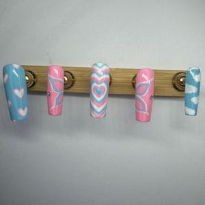 Pode incluir: Um conjunto de cinco unhas postiças longas e retangulares. As unhas apresentam vários desenhos, incluindo corações, asas de borboleta e padrões de nuvens, em tons de rosa, azul e branco. As unhas são montadas numa base de madeira com ferragens douradas.