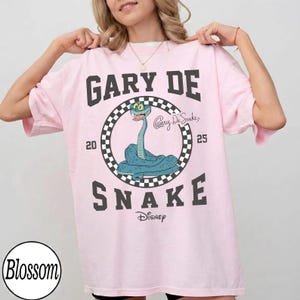 Puede incluir: Camiseta rosa claro con un dise&ntilde;o de serpiente de dibujos animados. El texto "GARY DE SNAKE" est&aacute; encima de la serpiente, con "20" y "25" a cada lado. La serpiente est&aacute; dentro de un c&iacute;rculo a cuadros. La palabra "Disney" est&aacute; debajo.