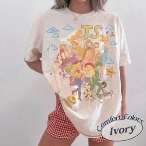 K&ouml;nnte beinhalten: Ein elfenbeinfarbenes Kurzarm-T-Shirt mit einem gro&szlig;en Grafikdruck von mehreren animierten Figuren in verschiedenen Outfits, darunter ein Sheriffstern, eine Rakete und die Buchstaben TS. Das Design enth&auml;lt Wolken und einen bunten Ball.