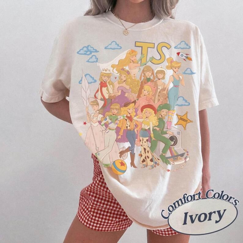 K&ouml;nnte beinhalten: Ein elfenbeinfarbenes Kurzarm-T-Shirt mit einem gro&szlig;en Grafikdruck von mehreren animierten Figuren in verschiedenen Outfits, darunter ein Sheriffstern, eine Rakete und die Buchstaben TS. Das Design enth&auml;lt Wolken und einen bunten Ball.