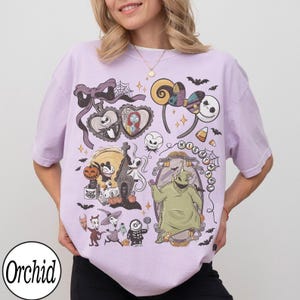 K&ouml;nnte beinhalten: Ein hellviolettes T-Shirt mit einem Grafikdruck von Charakteren und Motiven aus Nightmare Before Christmas, darunter Jack Skellington, Sally, Oogie Boogie, Zero sowie verschiedene Halloween-Themen wie Flederm&auml;use und K&uuml;rbisse.