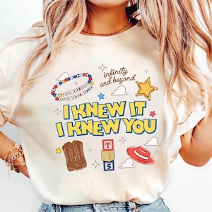 Puede incluir: Una camiseta color crema con un estampado gr&aacute;fico que incluye el texto "I KNEW IT I KNEW YOU" y "infinity and beyond". El dise&ntilde;o presenta una pulsera de cuentas, una estrella amarilla, botas vaqueras marrones, bloques de letras y un sombrero rojo.
