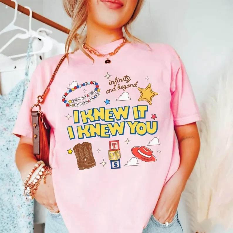 Puede incluir: Una camiseta rosa claro con un estampado gr&aacute;fico que incluye el texto "I KNEW IT I KNEW YOU", "infinity and beyond" y "YOU'VE GOT A FRIEND IN ME". El dise&ntilde;o presenta ilustraciones de una pulsera de cuentas, una estrella amarilla, botas vaqueras, bloques y un sombrero.