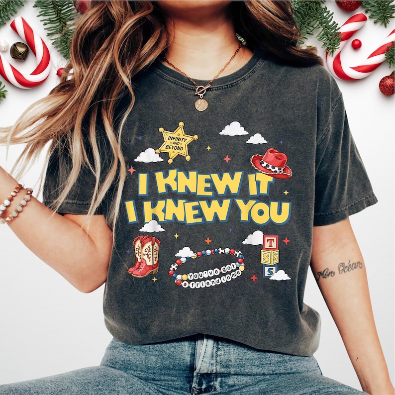 May include: A charcoal gray t-shirt featuring the yellow text I KNEW IT I KNEW YOU. Graphics include a gold sheriff star, a red cowboy hat, red cowboy boots, a beaded bracelet with the text YOU'VE GOT A FRIEND IN ME, and alphabet blocks.