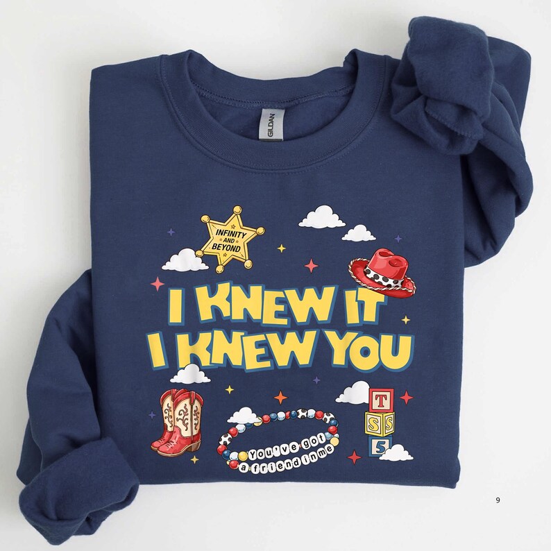 May include: A navy blue crewneck sweatshirt featuring a graphic print with the yellow text I KNEW IT I KNEW YOU, a yellow sheriff star badge, a red cowboy hat, red cowboy boots, a beaded bracelet, and stacked alphabet blocks with the letters T S 5.