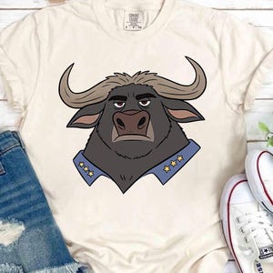 K&ouml;nnte beinhalten: Cremefarbenes T-Shirt mit einer Cartoon-Illustration eines B&uuml;ffels. Der B&uuml;ffel hat dunkles Fell, gebogene H&ouml;rner und einen blauen Kragen mit goldenen Sternen. Das Shirt wird mit Jeansshorts und wei&szlig;en Turnschuhen kombiniert.