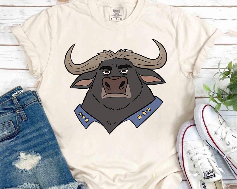 K&ouml;nnte beinhalten: Cremefarbenes T-Shirt mit einer Cartoon-Illustration eines B&uuml;ffels. Der B&uuml;ffel hat dunkles Fell, gebogene H&ouml;rner und einen blauen Kragen mit goldenen Sternen. Das Shirt wird mit Jeansshorts und wei&szlig;en Turnschuhen kombiniert.