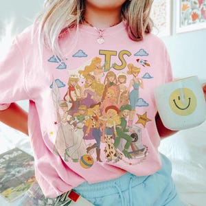 K&ouml;nnte beinhalten: Ein hellrosa T-Shirt mit einem Grafikdruck verschiedener animierter Figuren in unterschiedlichen Outfits, darunter ein Raumanzug, Cowgirl-Kleidung und Abendkleider. Das Design enth&auml;lt die Buchstaben TS, eine Rakete, Wolken und einen Stern.