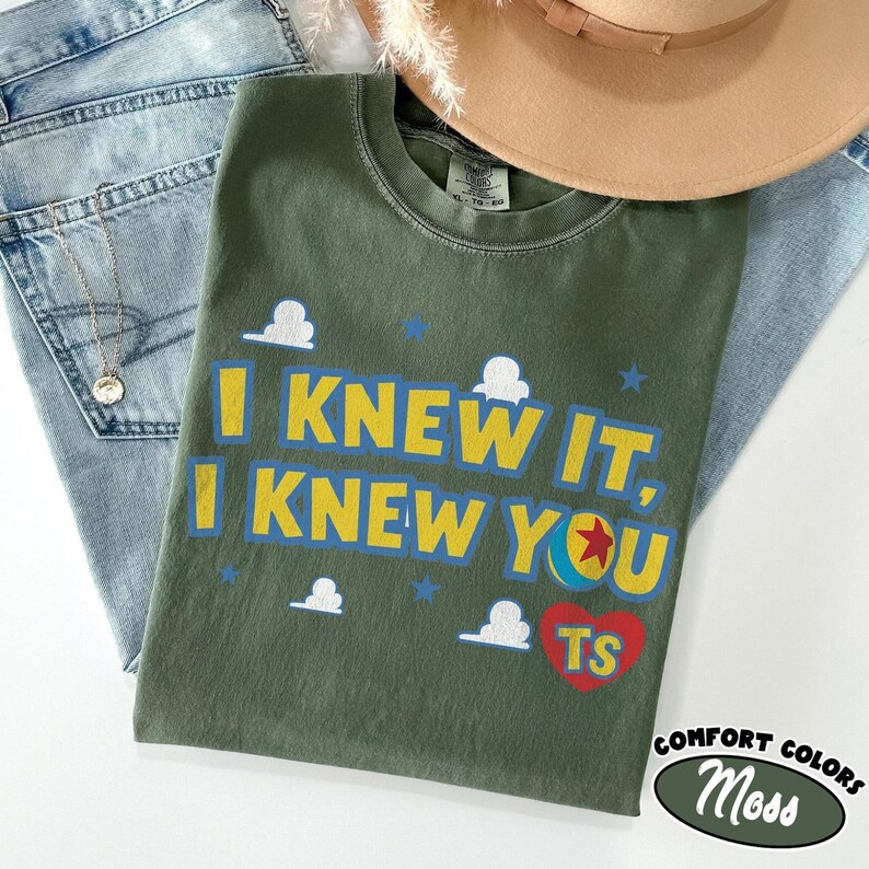 May include: An olive green crewneck t-shirt featuring the yellow text I KNEW IT, I KNEW YOU, a colorful ball graphic, a red heart with the letters TS, and small white cloud and blue star icons printed on the front. The shirt has a tag labeled XL-TG-EG.