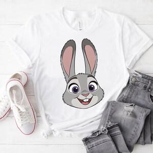 K&ouml;nnte beinhalten: Wei&szlig;es T-Shirt mit einem Cartoon-Kaninchengesicht in Grau, Rosa und Lila. Das Kaninchen l&auml;chelt. Das Shirt wird mit grauen Jeans und wei&szlig;en Turnschuhen kombiniert.