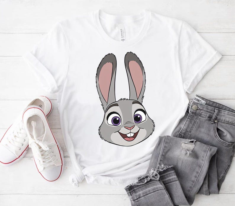 K&ouml;nnte beinhalten: Wei&szlig;es T-Shirt mit einem Cartoon-Kaninchengesicht in Grau, Rosa und Lila. Das Kaninchen l&auml;chelt. Das Shirt wird mit grauen Jeans und wei&szlig;en Turnschuhen kombiniert.