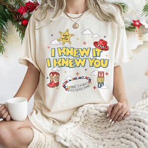 May include: A cream-colored t-shirt featuring the text "I KNEW IT I KNEW YOU" in yellow with blue outlines. Graphics include a yellow sheriff star, a red cowboy hat, red cowboy boots, a beaded bracelet with "YOU'VE GOT A FRIEND IN ME" text, and blocks.