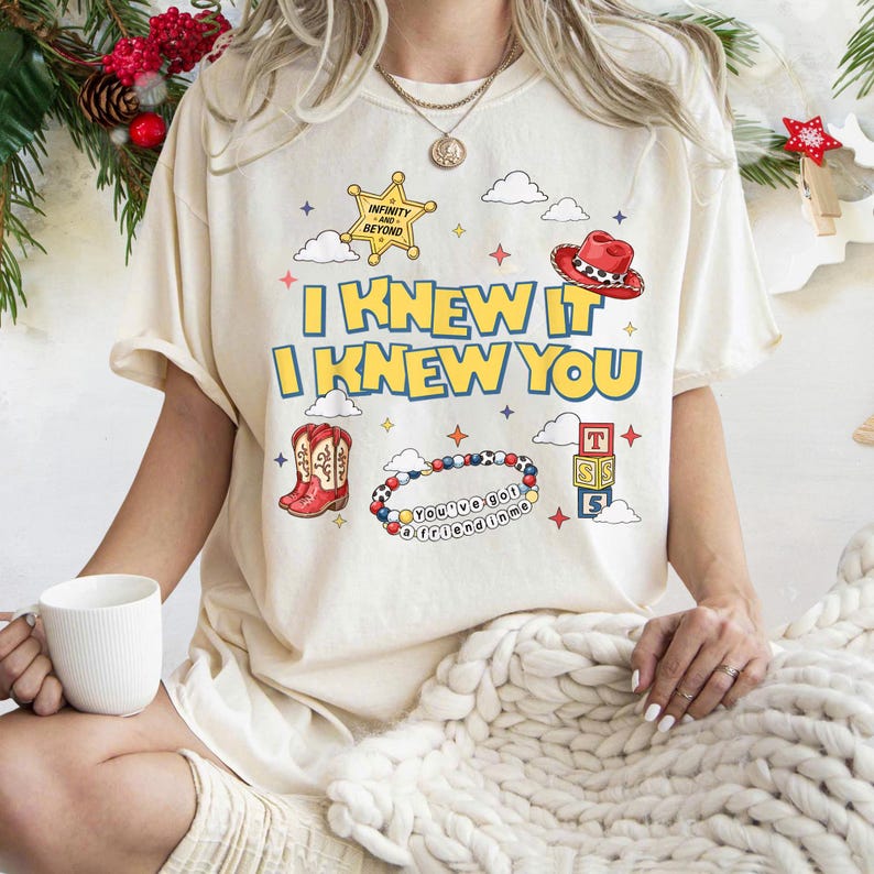 May include: A cream-colored t-shirt featuring the text "I KNEW IT I KNEW YOU" in yellow with blue outlines. Graphics include a yellow sheriff star, a red cowboy hat, red cowboy boots, a beaded bracelet with "YOU'VE GOT A FRIEND IN ME" text, and blocks.