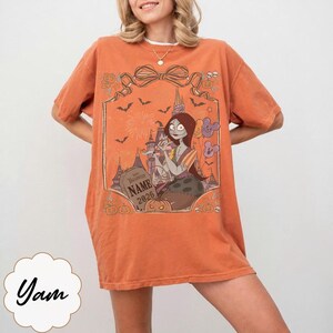 May include: An orange short-sleeve t-shirt featuring a graphic print of a female character with long red hair, a castle, bats, and pumpkins. The design includes a tombstone with the text "HAPPY HALLOWEEN NAME 2026" and Mickey-shaped balloons.