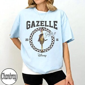 Puede incluir: Camiseta azul claro con la palabra "GAZELLE" en letras oscuras sobre un gr&aacute;fico circular. El gr&aacute;fico incluye un personaje de gacela de dibujos animados, los a&ntilde;os "20" y "16" y el logotipo de Disney. La camiseta es de estilo casual.