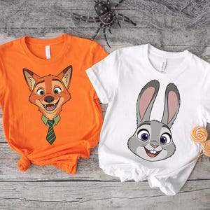 K&ouml;nnte beinhalten: Zwei kurz&auml;rmelige T-Shirts. Eines ist orange mit einem Cartoon-Fuchsgesicht mit gr&uuml;ner Krawatte. Das andere ist wei&szlig; mit einem Cartoon-Kaninchengesicht. Beide Shirts befinden sich auf einer verwitterten Holzoberfl&auml;che.