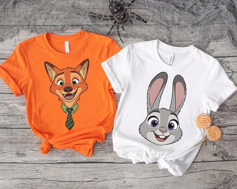 K&ouml;nnte beinhalten: Zwei kurz&auml;rmelige T-Shirts. Eines ist orange mit einem Cartoon-Fuchsgesicht mit gr&uuml;ner Krawatte. Das andere ist wei&szlig; mit einem Cartoon-Kaninchengesicht. Beide Shirts befinden sich auf einer verwitterten Holzoberfl&auml;che.