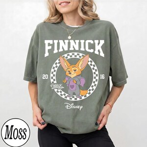 Puede incluir: Camiseta verde salvia con la palabra "FINNICK" en blanco sobre un personaje de zorro de dibujos animados. El personaje lleva un atuendo morado y sostiene un coraz&oacute;n. El logotipo de Disney est&aacute; en la parte inferior.